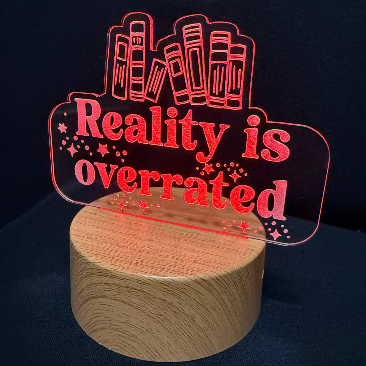A red laser-cut acrylic sign reading 'Reality is overrated' with book illustrations, mounted on a round wooden base against a dark navy background.
