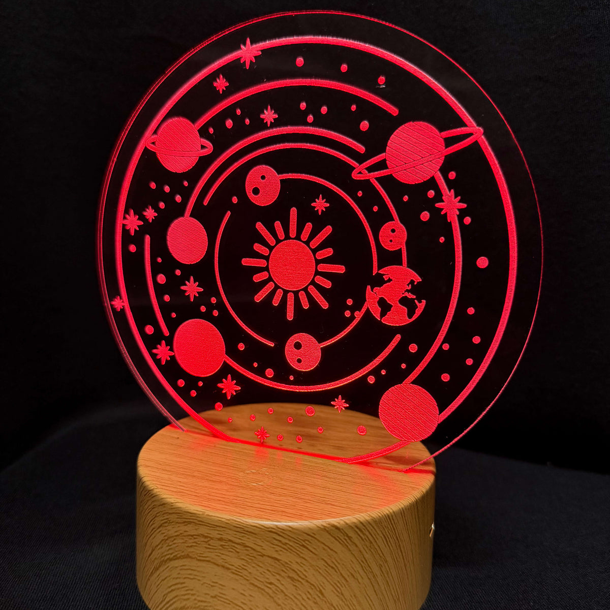A red laser-cut acrylic solar system night light on a round wooden base, set against a dark black background.