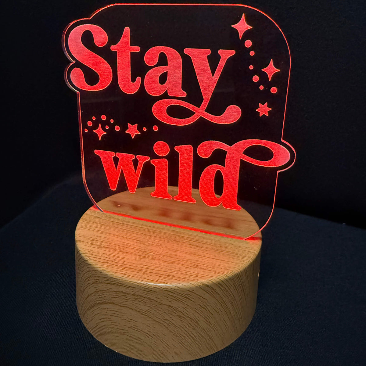 A red acrylic LED night light with the words 'Stay wild' in a stylized font, mounted on a round wooden base against a dark background.