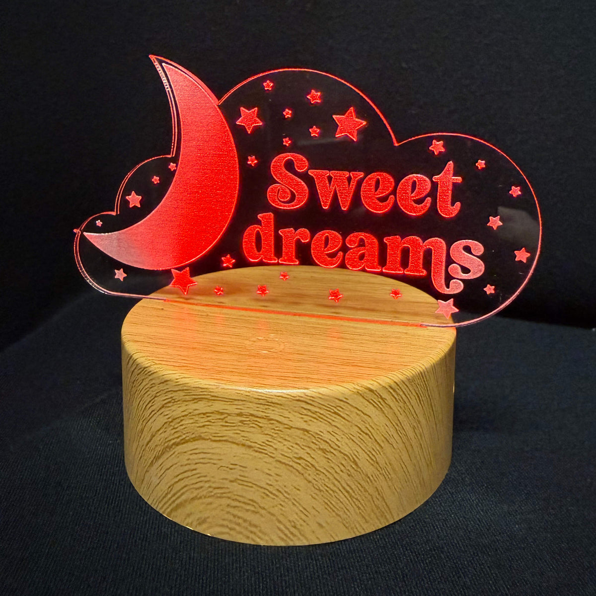 A decorative acrylic night light or wall sign shaped like a crescent moon and cloud with the words 'Sweet dreams' in red, mounted on a cylindrical wooden base against a black background.