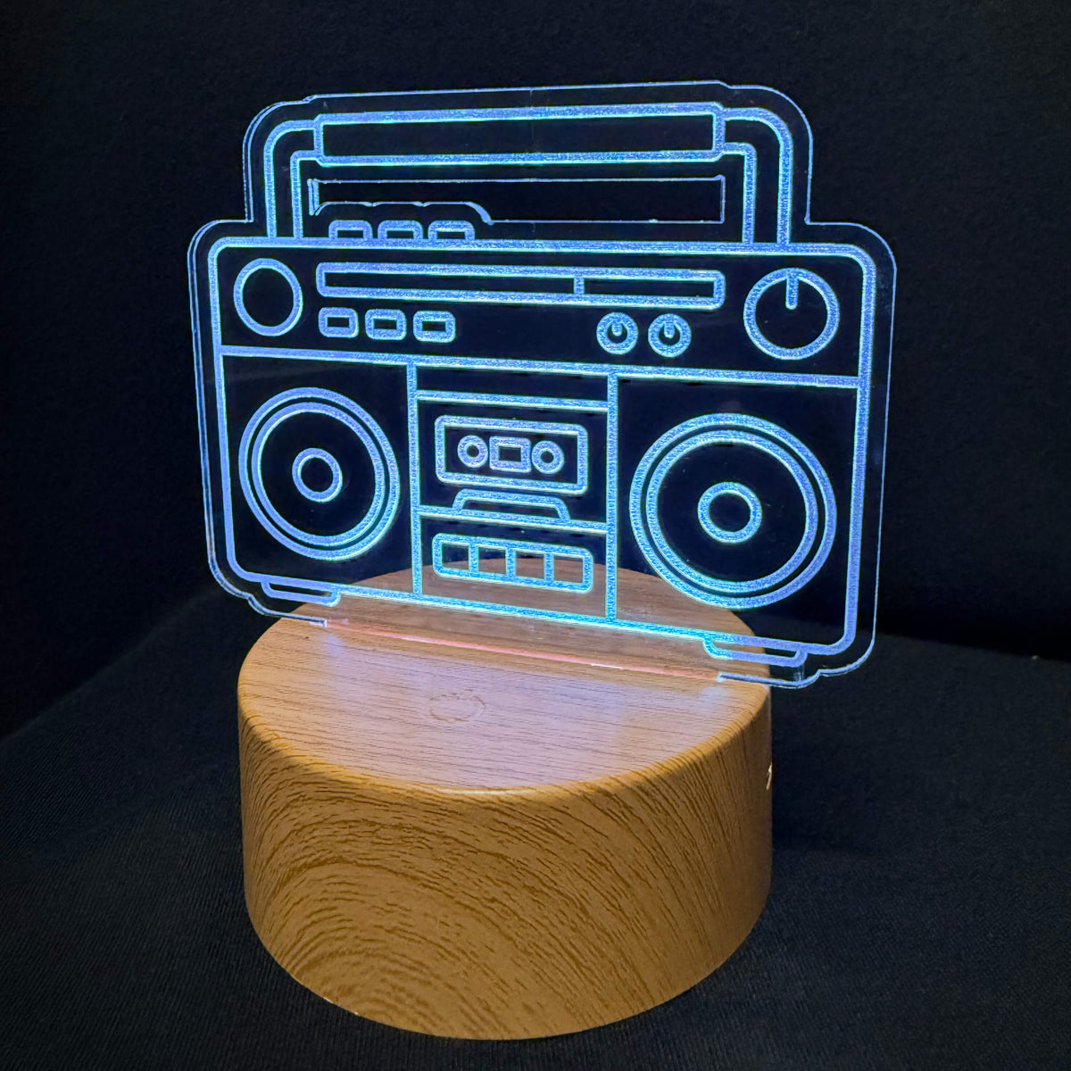 An LED acrylic lamp featuring a glowing blue line-art design of a vintage boombox on top of a cylindrical light-wood base, set against a solid black background.
