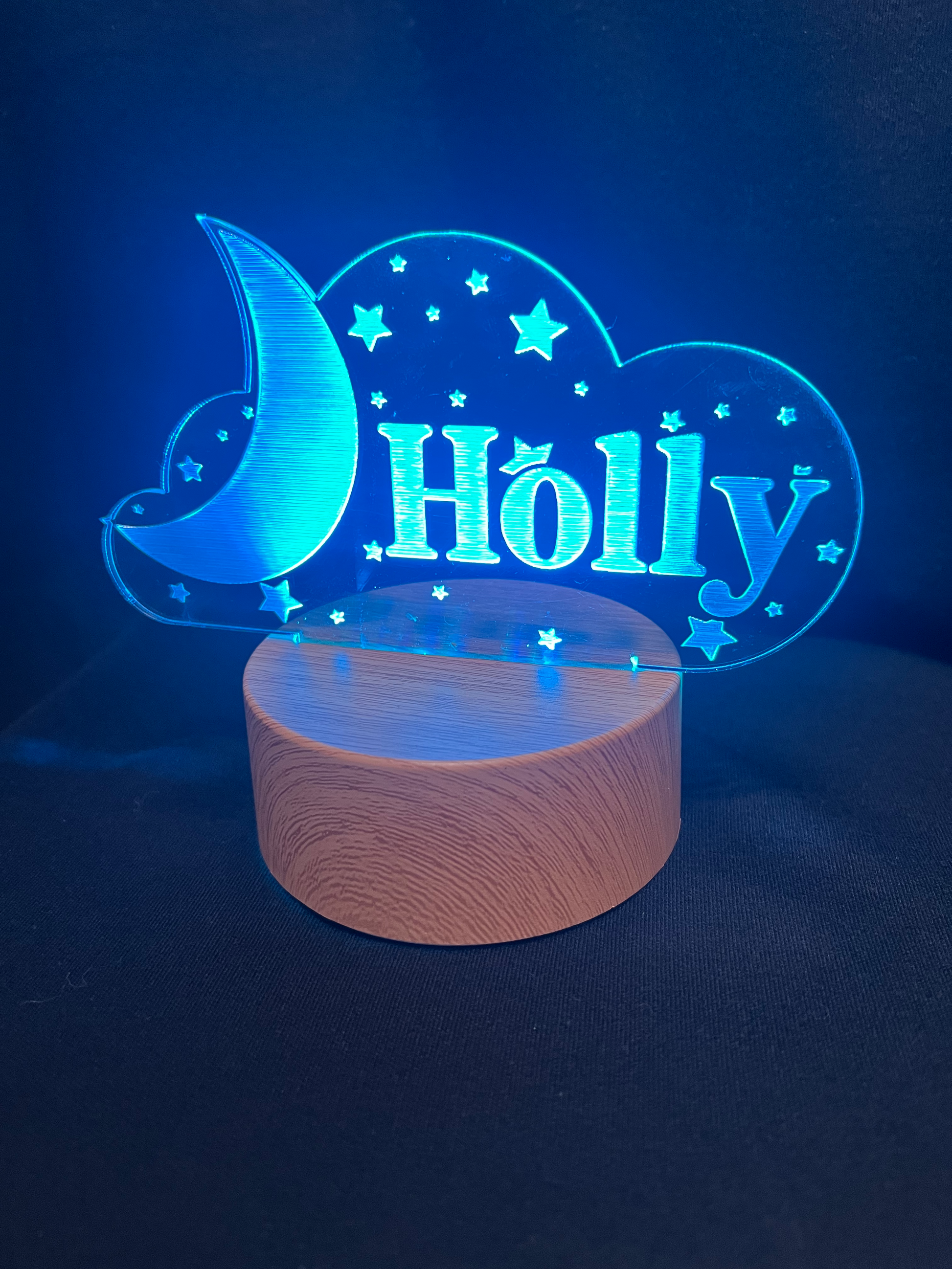 Custom acrylic LED night light with the name 'Holly' engraved in a cloud and moon design, glowing blue on a wooden-style base against a dark background