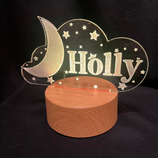 Custom acrylic LED night light with the name 'Holly' engraved in a cloud and moon design, glowing blue on a wooden-style base against a dark background