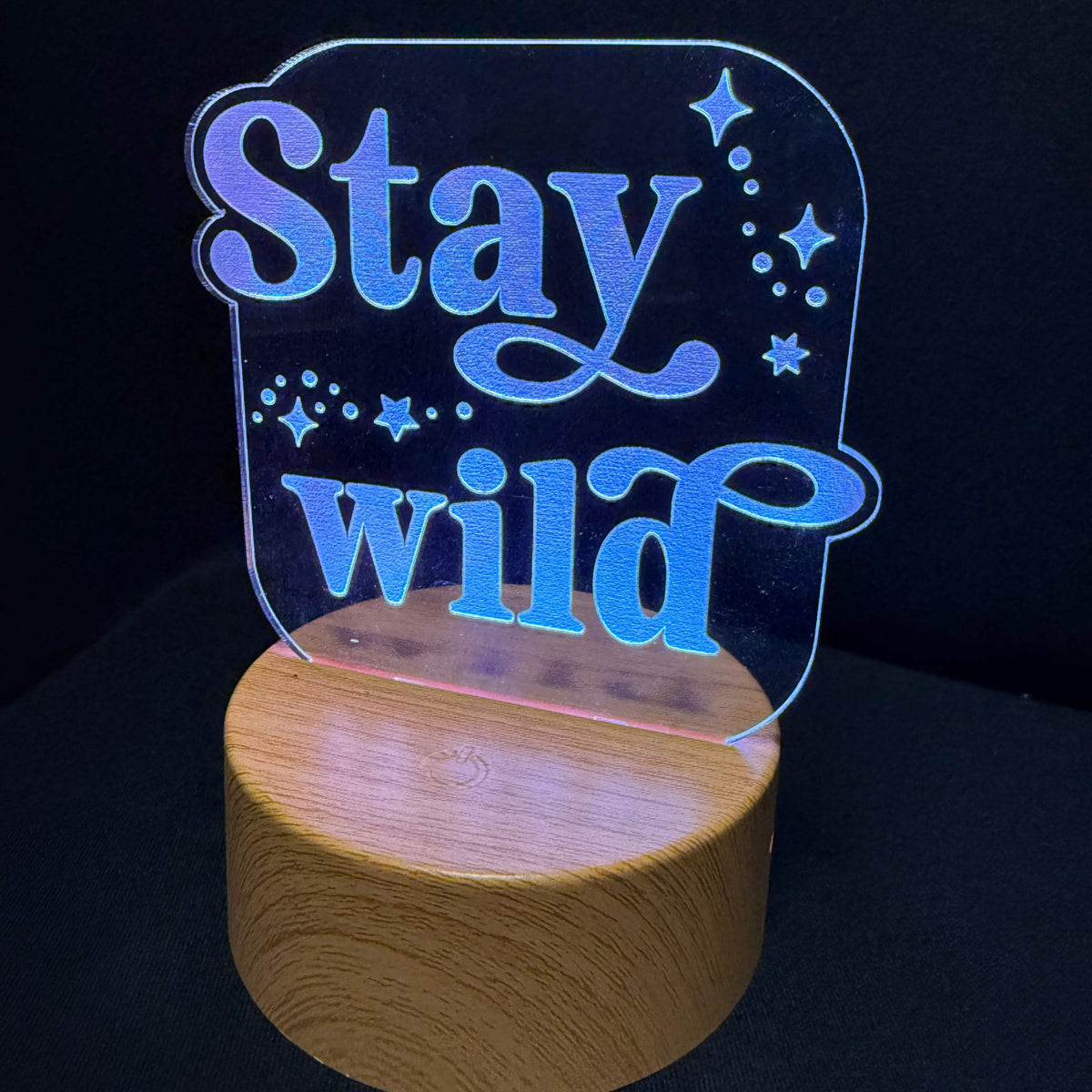 An LED acrylic night light with the phrase 'Stay wild' in glowing blue script, mounted on a round natural wood base against a black background.
