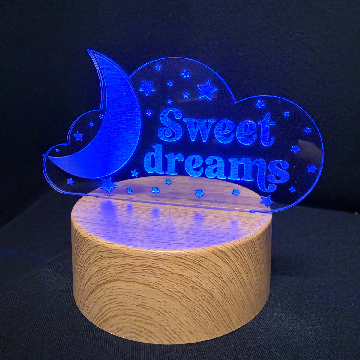 A wooden cylindrical lamp base with a clear acrylic nightstand sign that reads 'Sweet dreams' in blue, featuring a crescent moon and stars, set against a dark background.