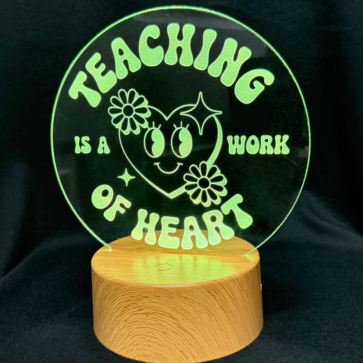 A circular acrylic LED night light lamp featuring the text 'TEACHING IS A WORK OF HEART' in a light green neon glow, mounted on a round wooden base and photographed against a black fabric background.