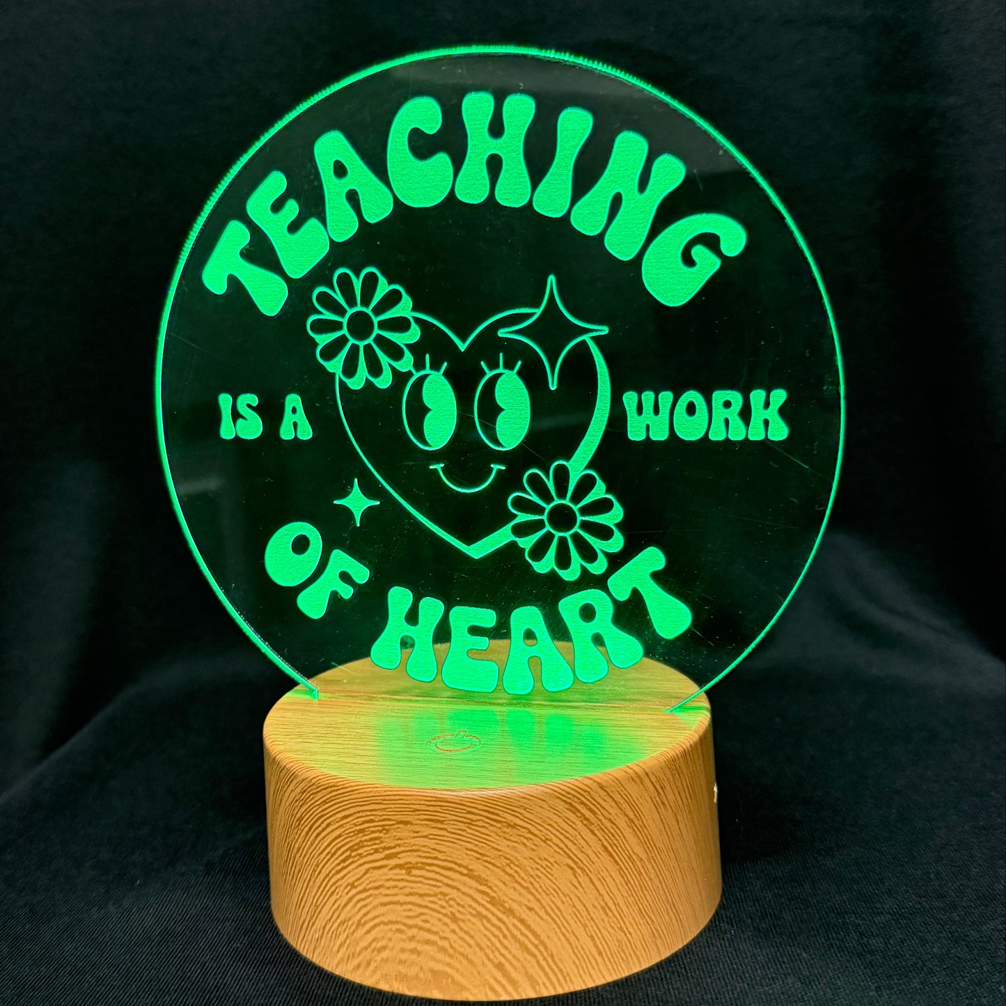 A circular acrylic LED night light on a cylindrical wooden base, glowing green with the text 'TEACHING IS A WORK OF HEART' and a smiling heart illustration, set against a black fabric background.