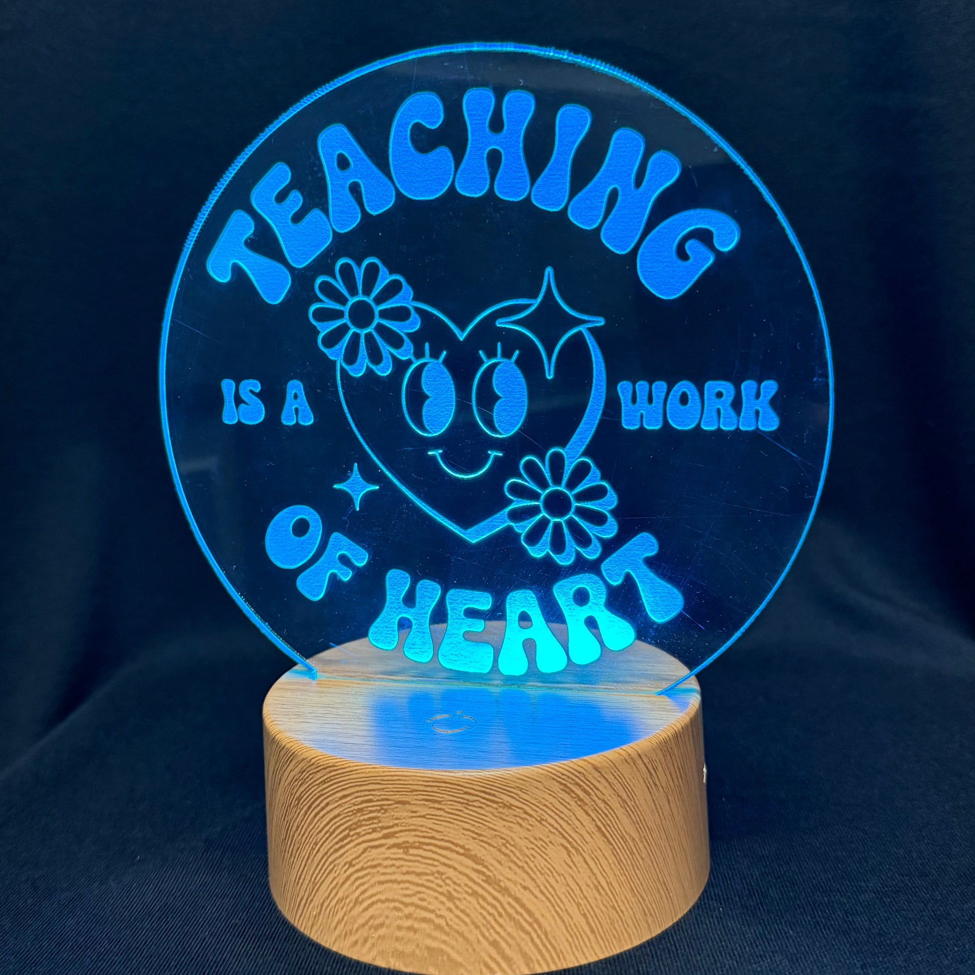 A circular acrylic LED lamp displaying the phrase 'TEACHING IS A WORK OF HEART' in glowing blue lettering around a smiling heart and flower design, mounted on a cylindrical natural wood base against a dark fabric background.