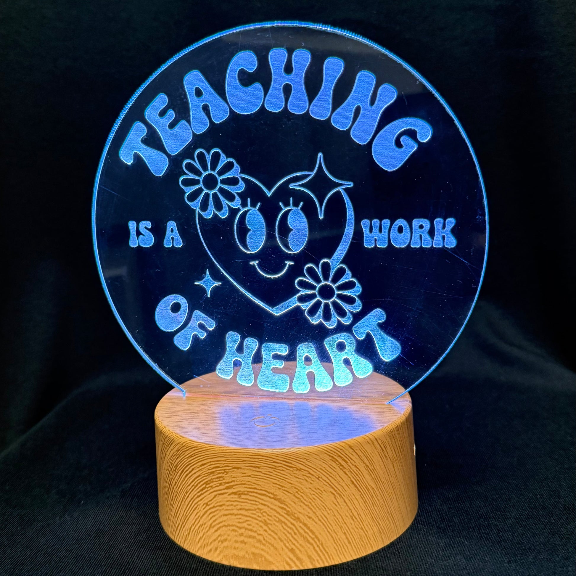 A circular acrylic LED night light on a wooden base, displaying the message 'TEACHING IS A WORK OF HEART' in glowing blue lettering against a black background.