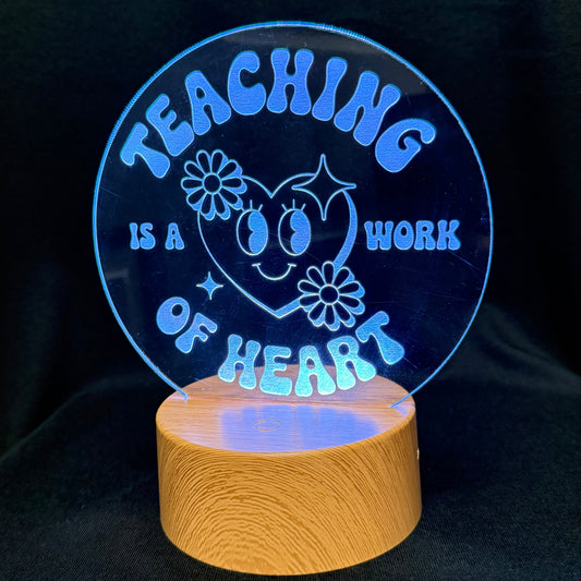 A circular acrylic LED night light on a wooden base, displaying the message 'TEACHING IS A WORK OF HEART' in glowing blue lettering against a black background.