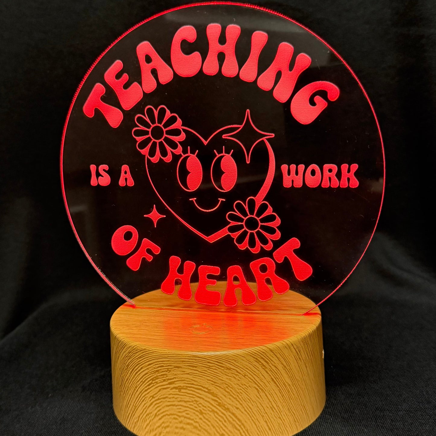 A circular red acrylic plaque with the text 'TEACHING IS A WORK OF HEART' and a smiling heart illustration, mounted on a round light wood base against a black fabric background.