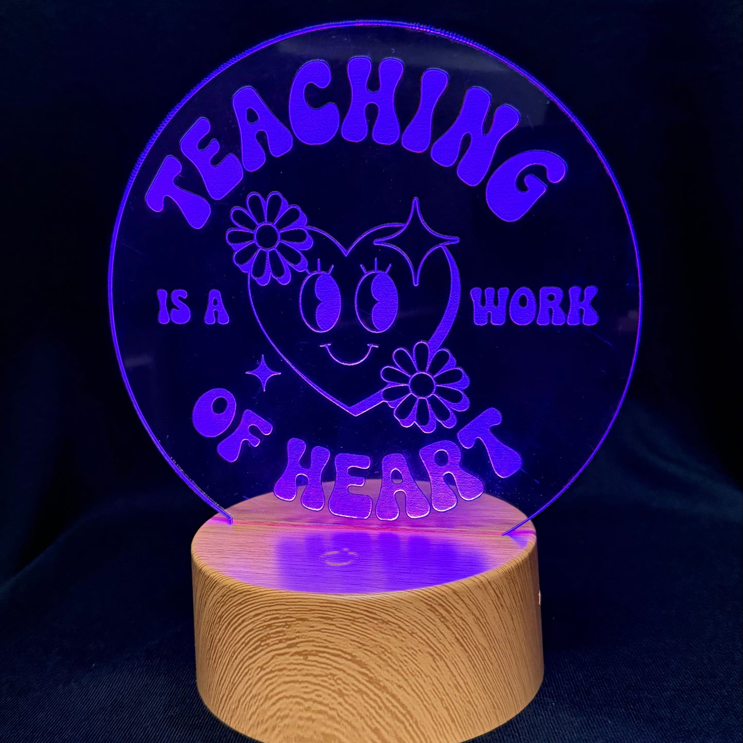 A circular acrylic LED night light on a wooden base, glowing with purple light that reads 'TEACHING IS A WORK OF HEART' with a smiling heart and flowers, set against a dark background.