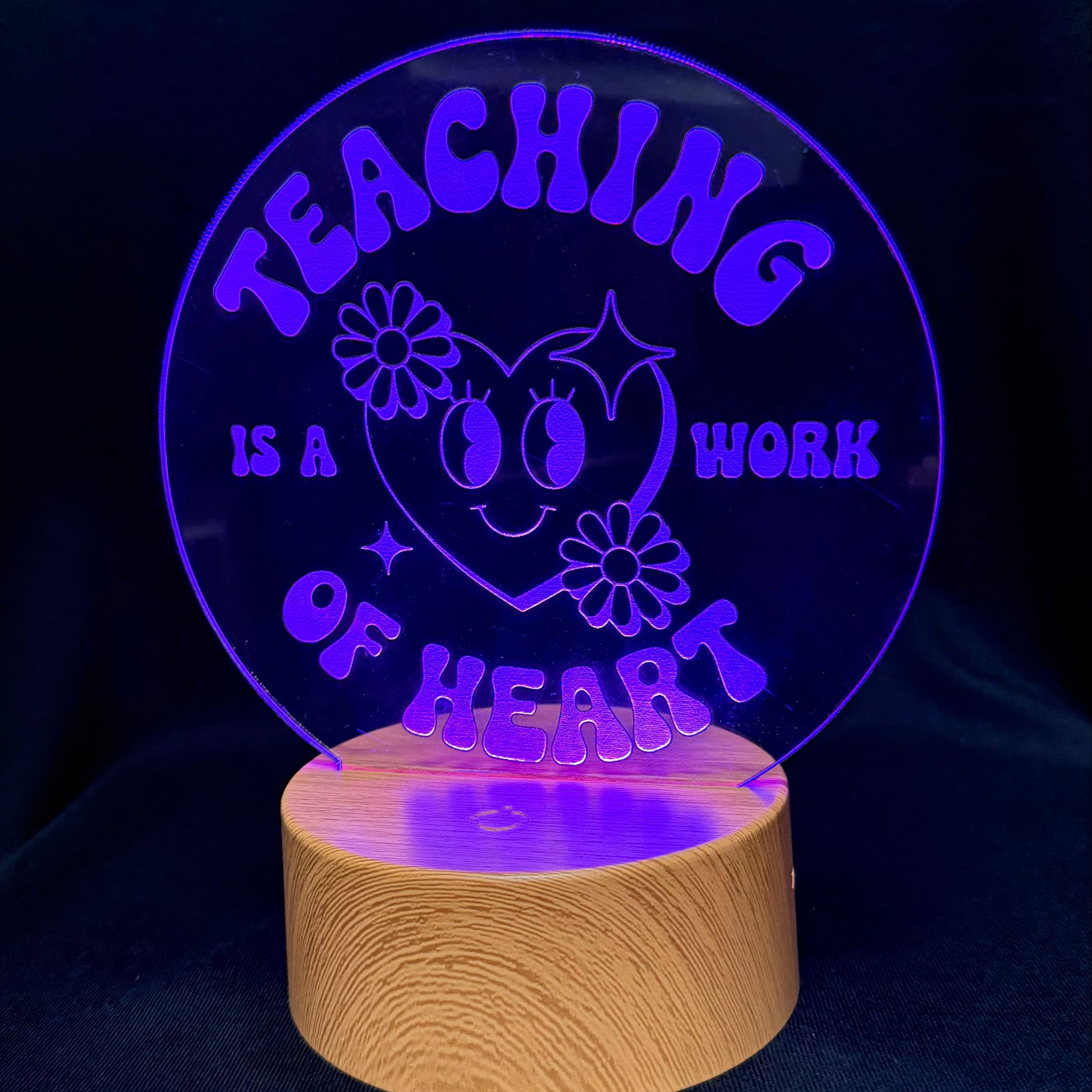 A circular acrylic LED night light on a wooden base, glowing with purple light that reads 'TEACHING IS A WORK OF HEART' with a smiling heart and flowers, set against a dark background.