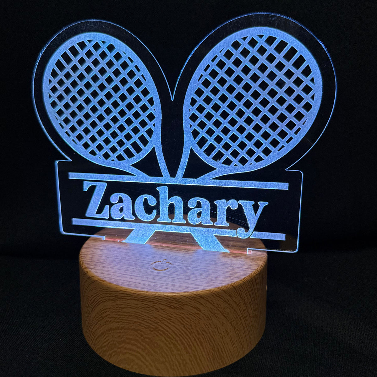 A personalized LED acrylic lamp featuring two tennis rackets and the name 'Zachary' illuminated in blue on a round wooden base, set against a black background.