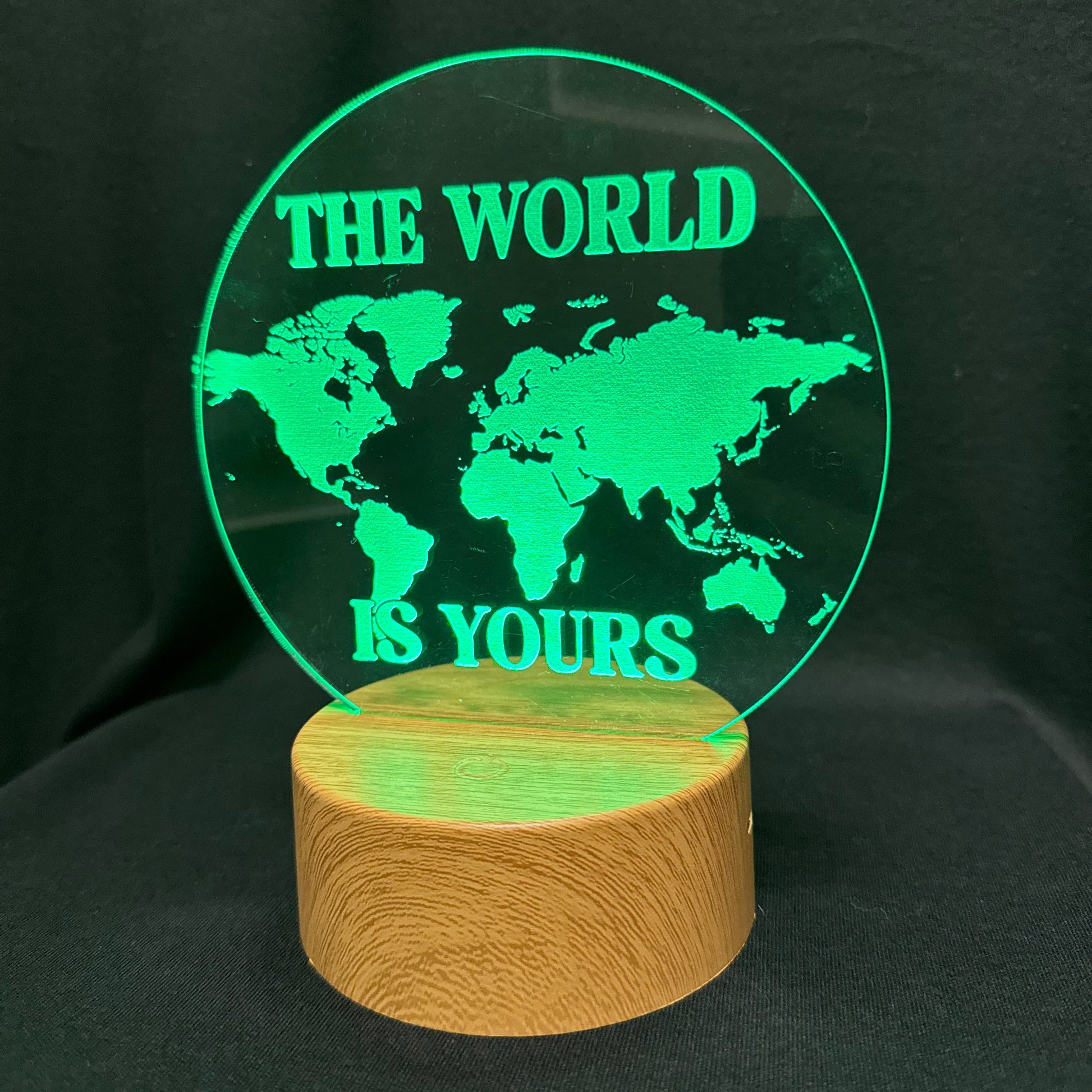 A circular clear acrylic lamp featuring a green illuminated world map design with the text 'THE WORLD IS YOURS' in green, mounted on a cylindrical natural wood base against a black background.