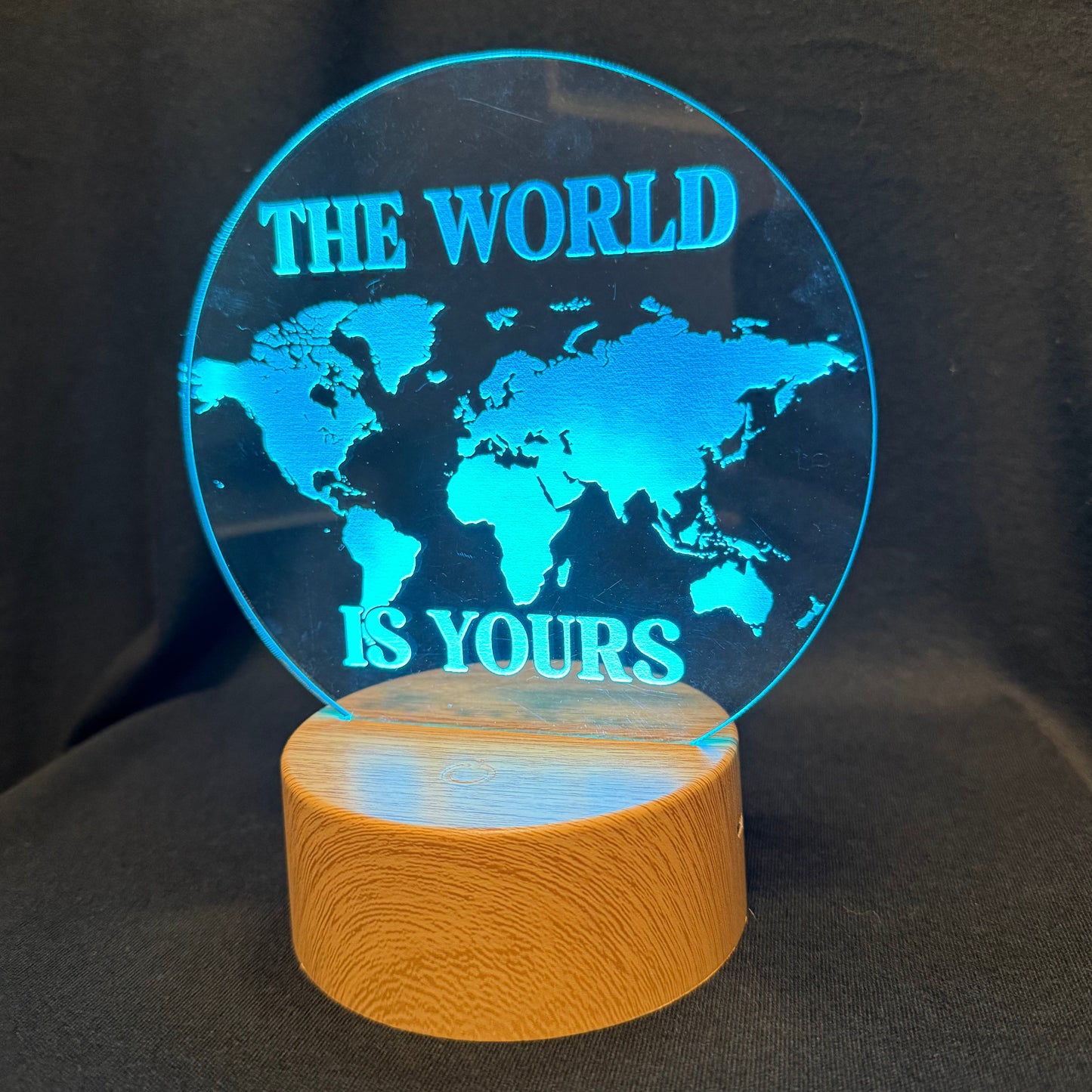 A circular acrylic LED lamp featuring a glowing blue world map design with the text 'THE WORLD IS YOURS' on a cylindrical wooden base, set against a dark fabric background.