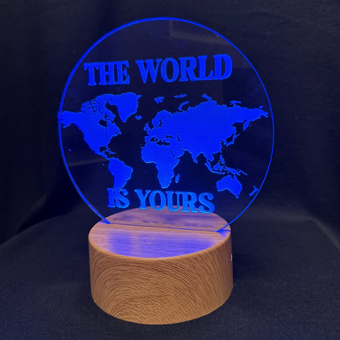 A circular acrylic LED lamp with a world map design and the text 'THE WORLD IS YOURS' in bright blue, mounted on a cylindrical wooden base against a dark background.