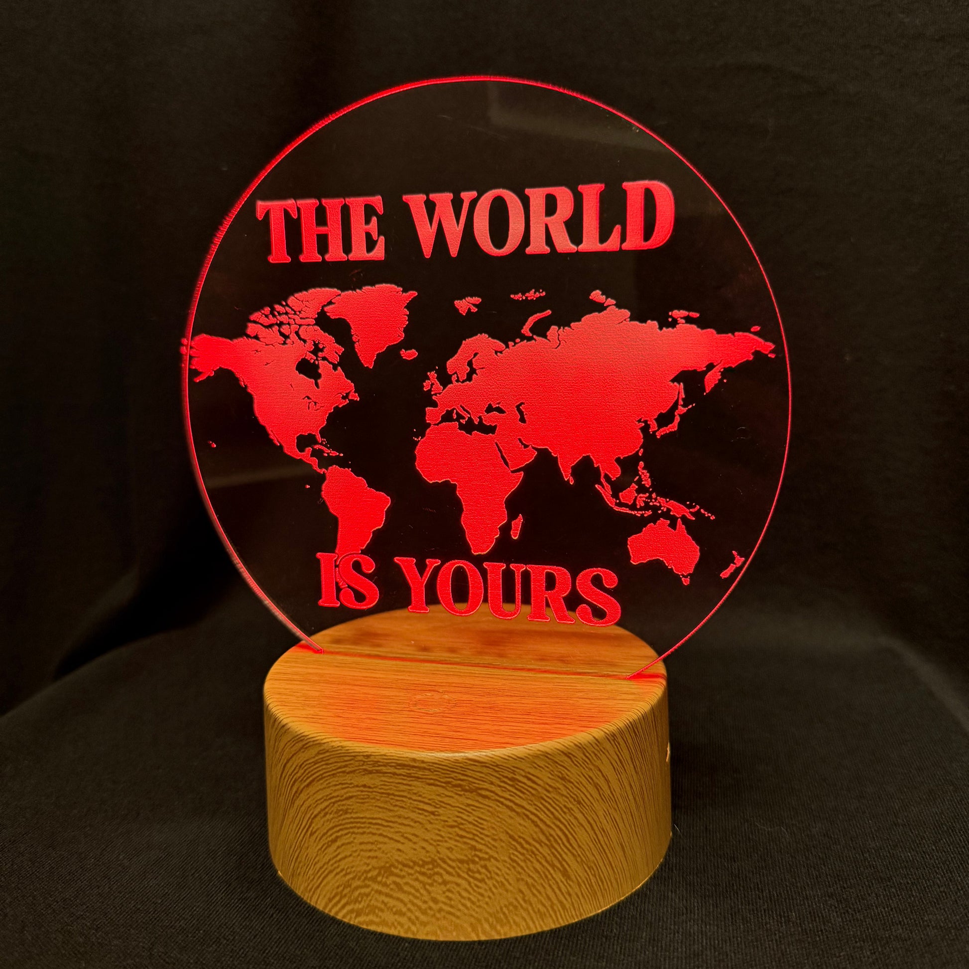 A circular acrylic LED light sign with a red world map design and the text 'THE WORLD IS YOURS' in red, mounted on a cylindrical wooden base against a black background.