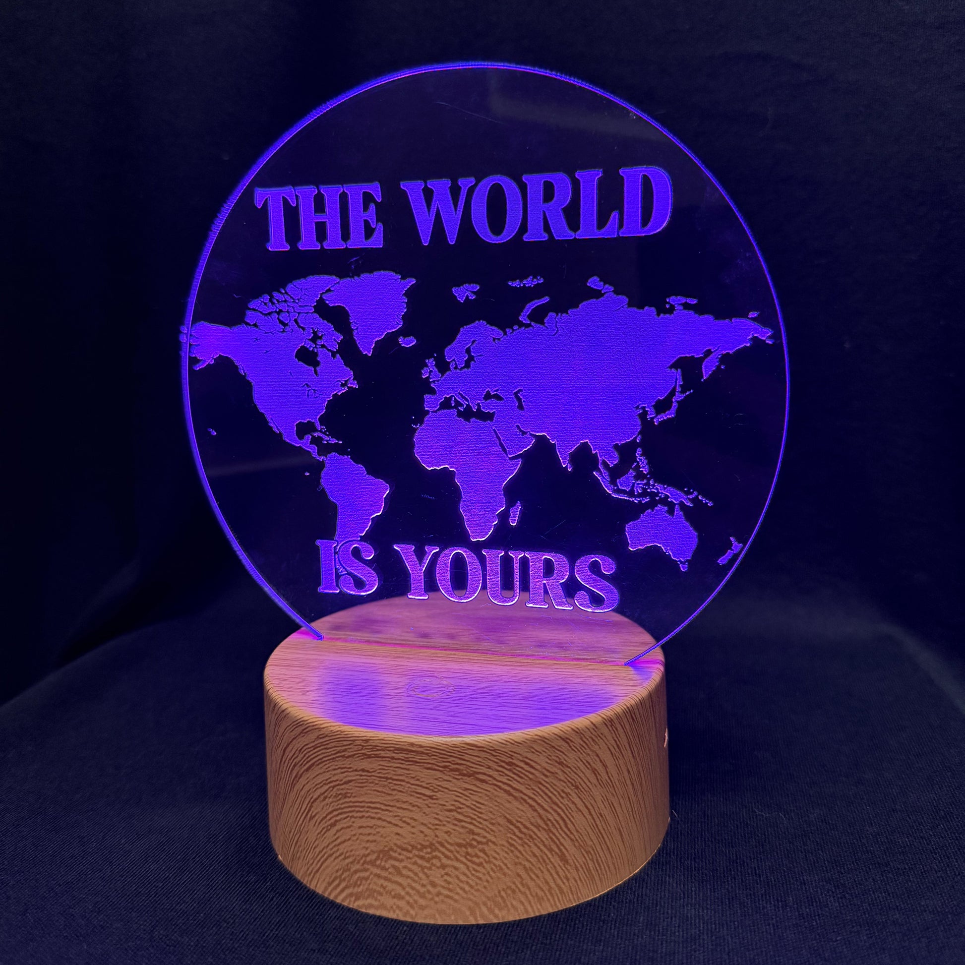 A circular acrylic LED night light with a world map design and the text 'THE WORLD IS YOURS' in purple, resting on a cylindrical wooden base against a dark background.