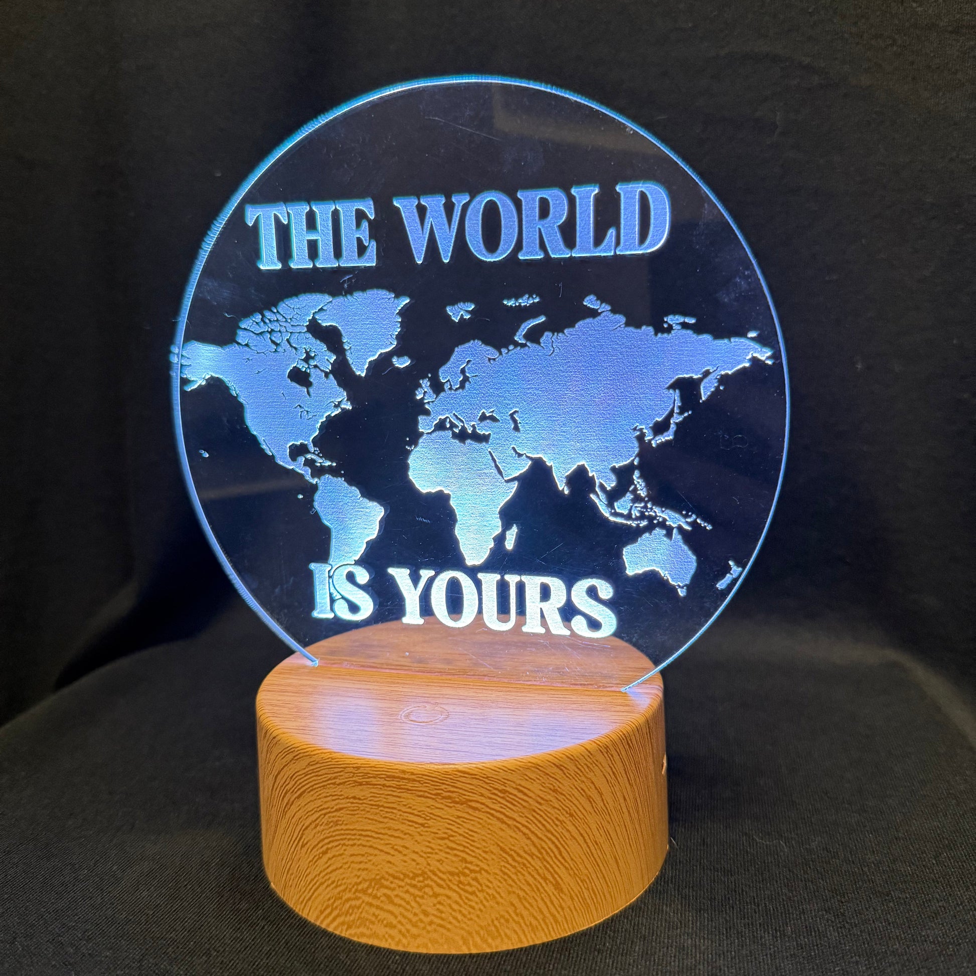 An illuminated acrylic world map lamp with the text 'THE WORLD IS YOURS' in white, mounted on a cylindrical wooden base against a black background.