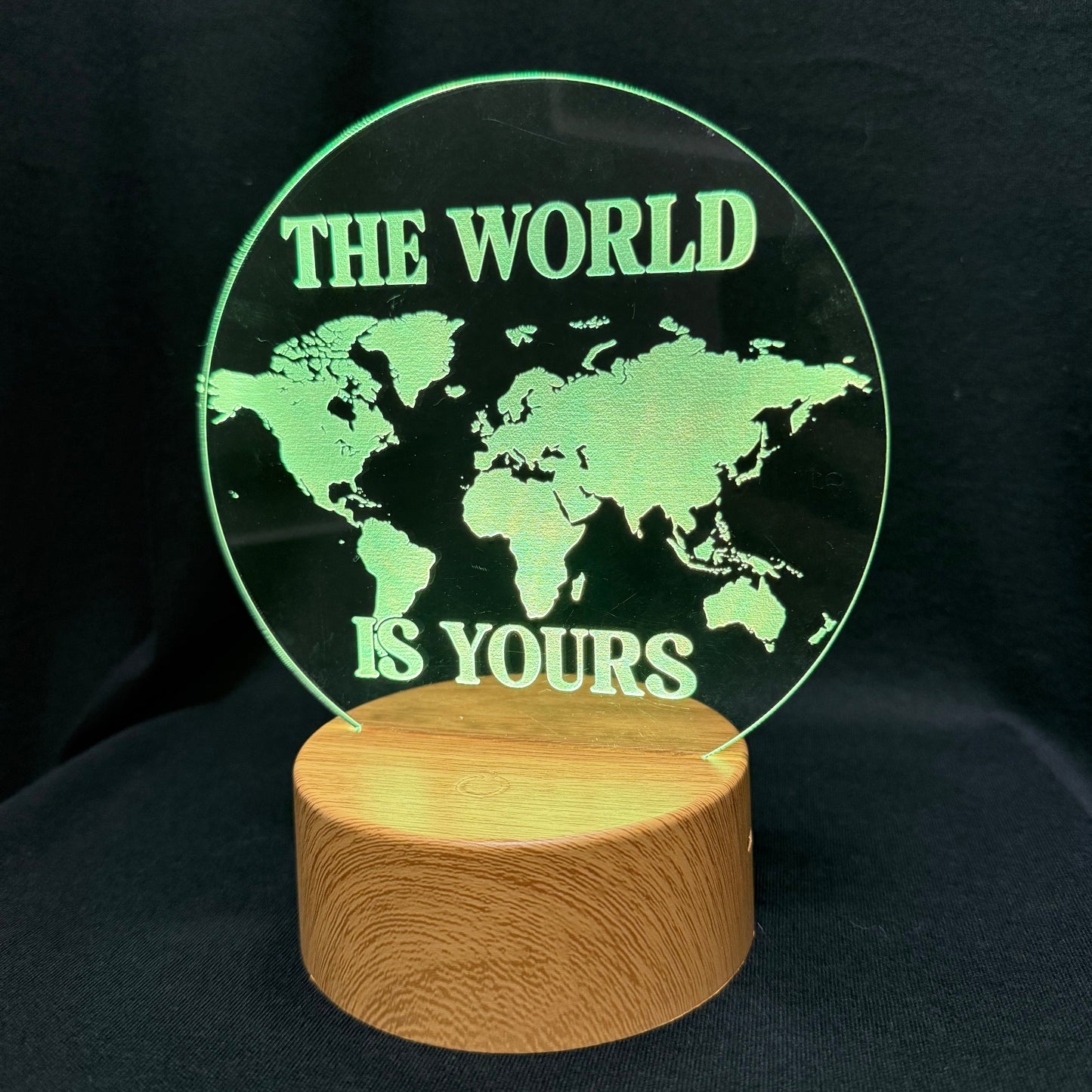 A circular clear acrylic lamp featuring a green world map design and the text 'THE WORLD IS YOURS' in green, mounted on a cylindrical light wood base against a black background.