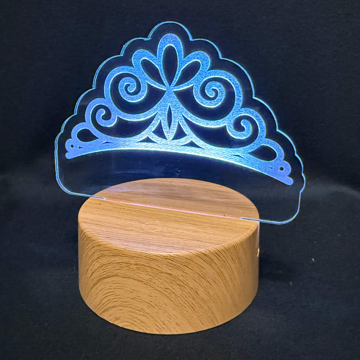 A 3D LED acrylic lamp featuring an ornate tiara design that glows with a blue light, mounted on a round wooden base against a black background.