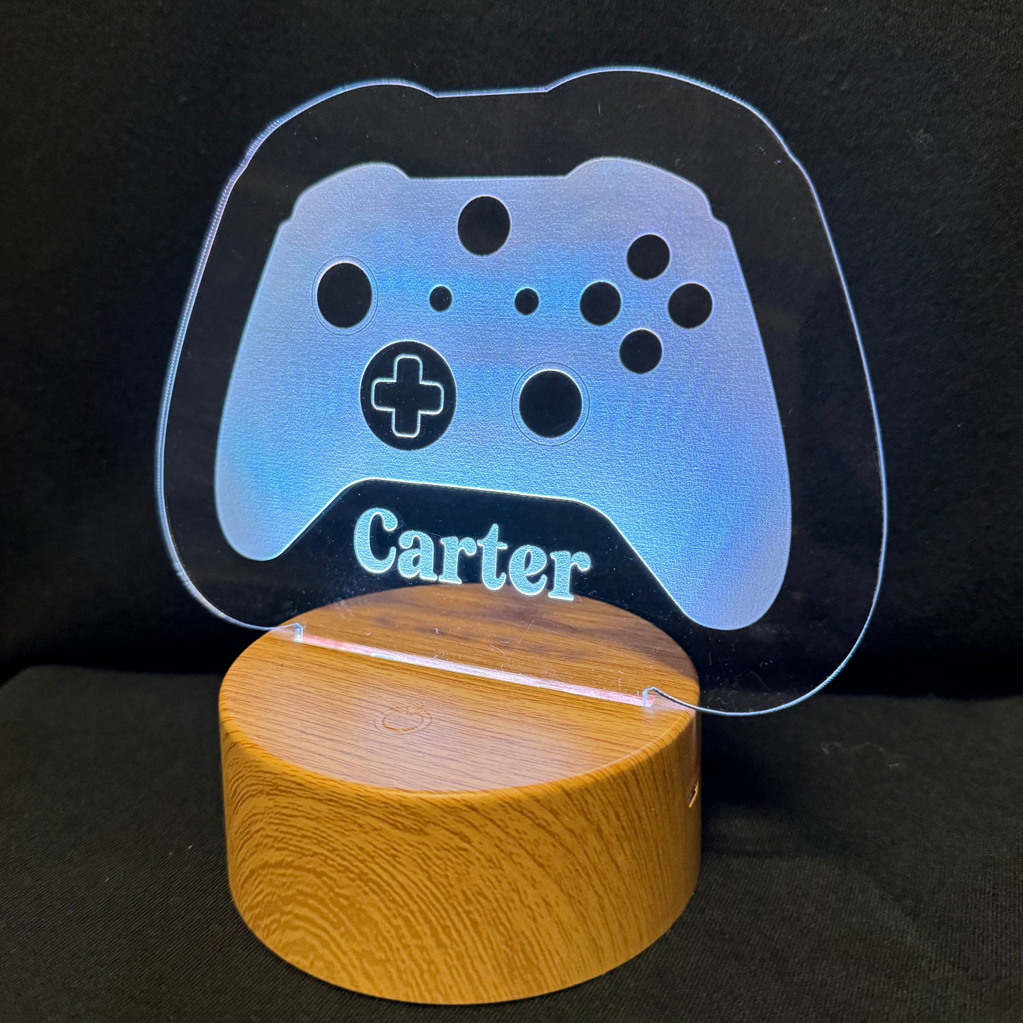 A personalized gaming-themed LED light featuring a glowing blue acrylic game controller silhouette with the name 'Carter' engraved, resting on a cylindrical wooden base against a dark background.