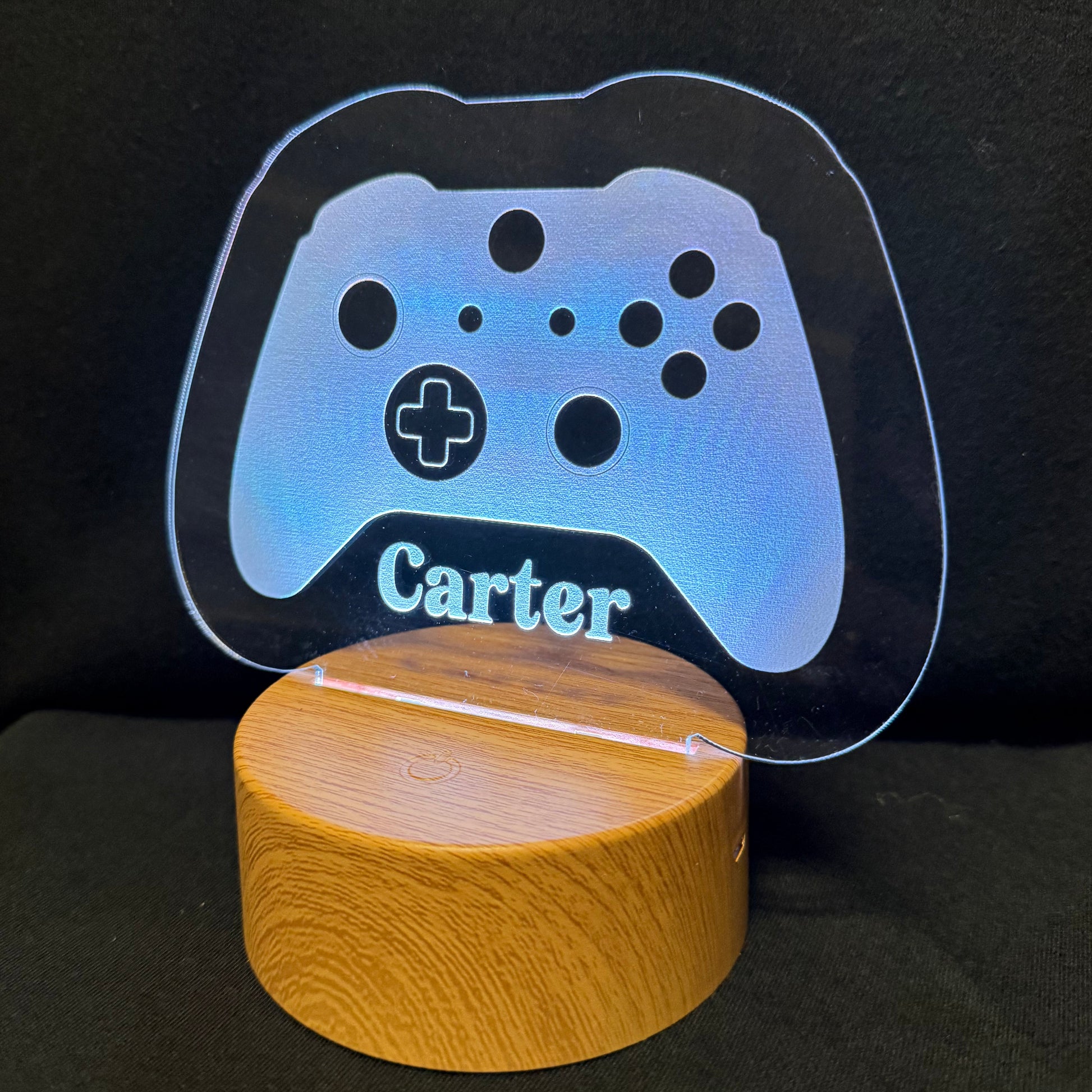 A personalized gaming-themed LED light featuring a glowing blue acrylic game controller silhouette with the name 'Carter' engraved, resting on a cylindrical wooden base against a dark background.