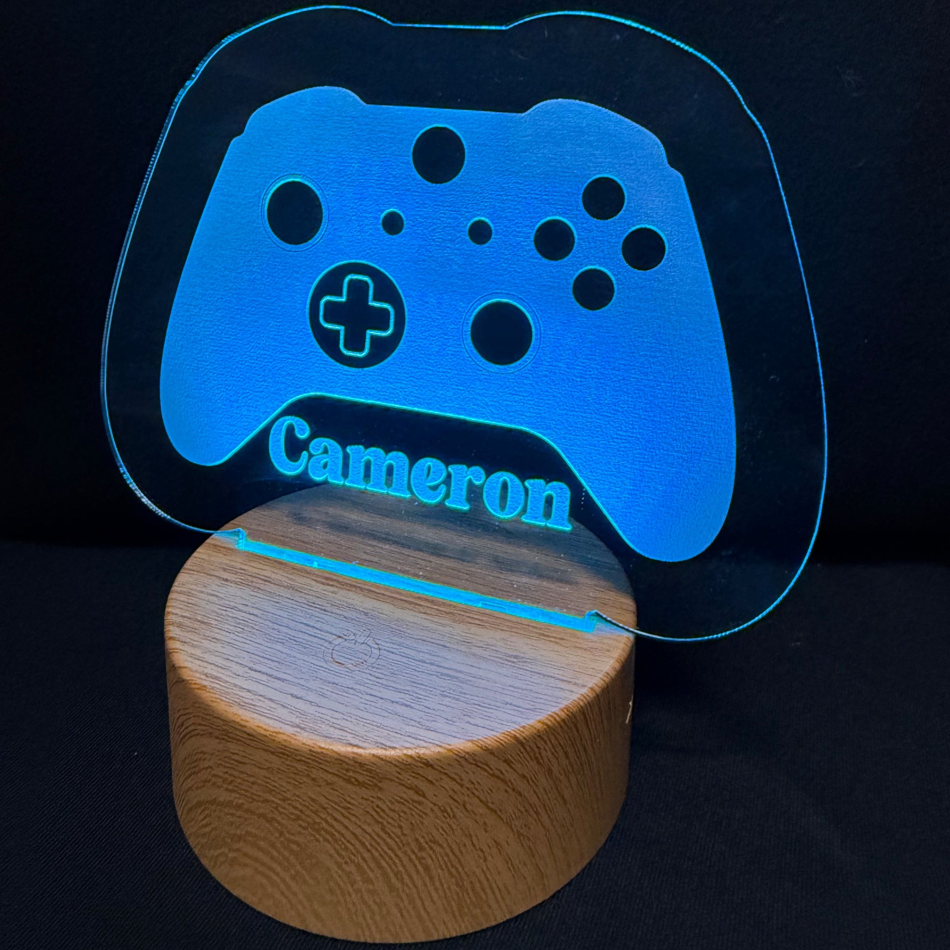 A personalized gaming controller-shaped LED light on a cylindrical wooden base, featuring the name 'Cameron' in blue on a black background.