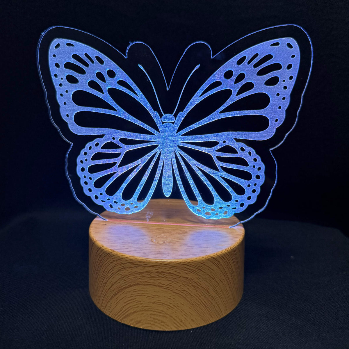 A butterfly-shaped LED night light with a glowing blue and purple acrylic body resting on a round wooden base, set against a dark background.