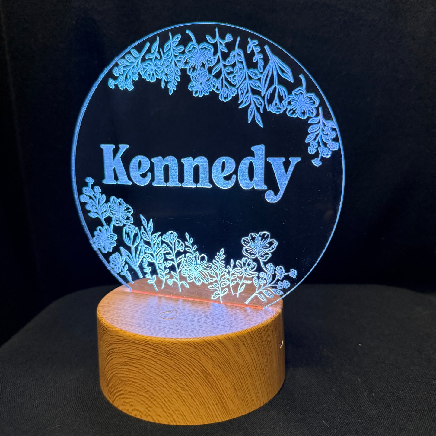 Personalized acrylic LED lamp with a circular glowing panel featuring the name 'Kennedy' in blue script, surrounded by floral designs, mounted on a cylindrical wooden base against a black background.
