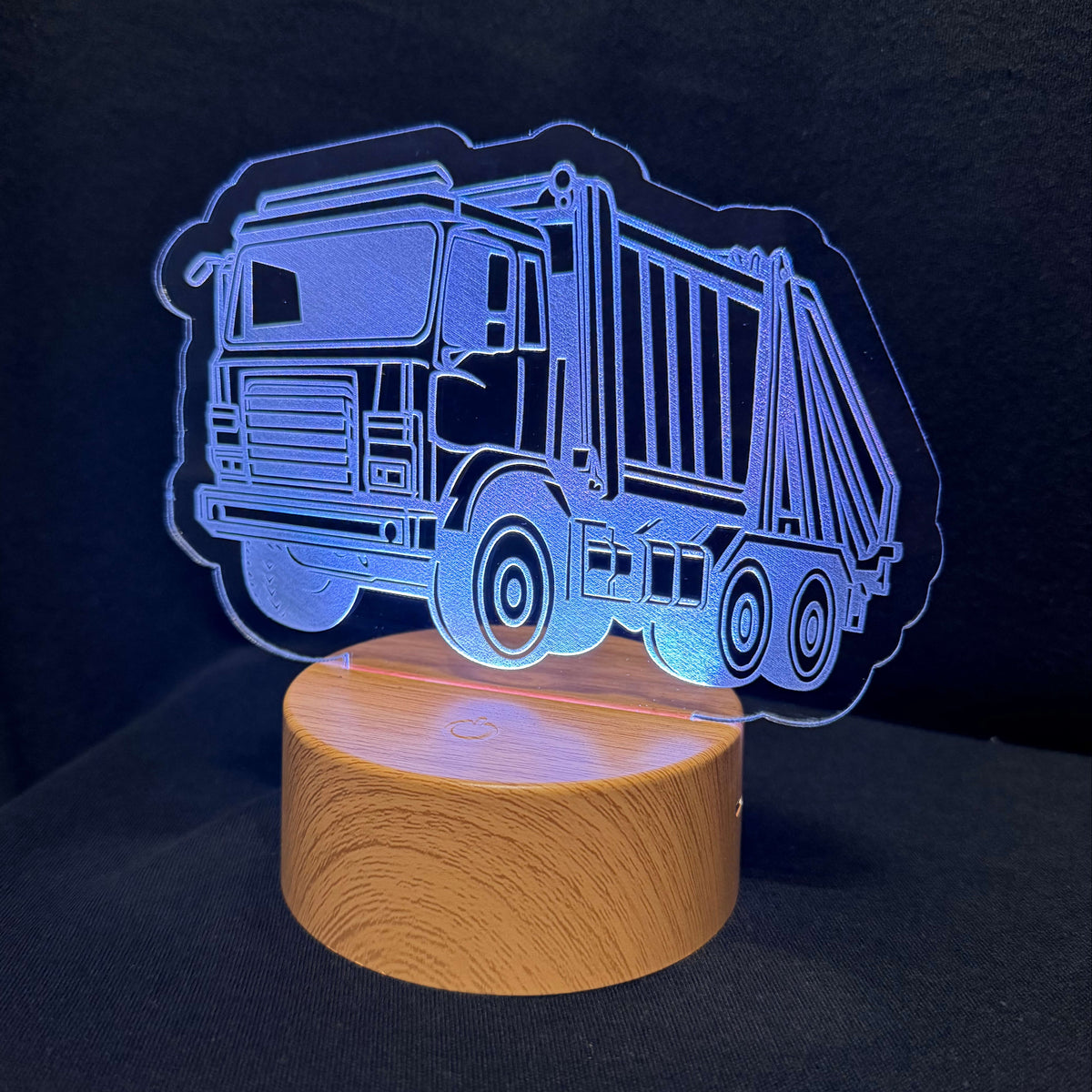 An LED acrylic night light featuring a glowing blue garbage truck silhouette on a round wooden base, set against a dark background.