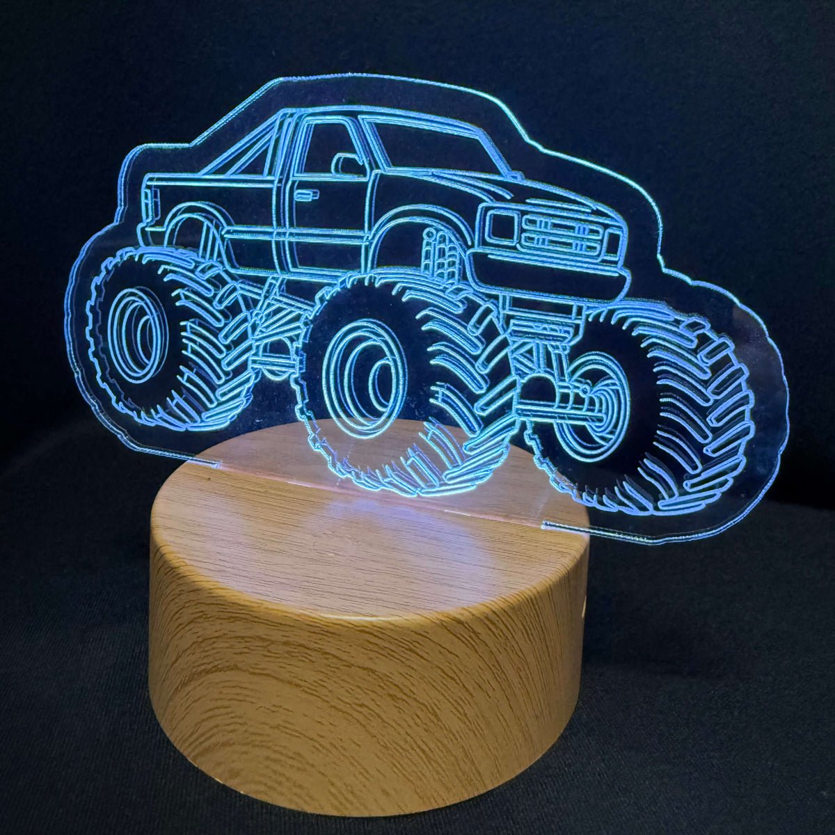 An LED acrylic night light featuring a glowing blue outline of a monster truck on a cylindrical wooden base, set against a dark background.