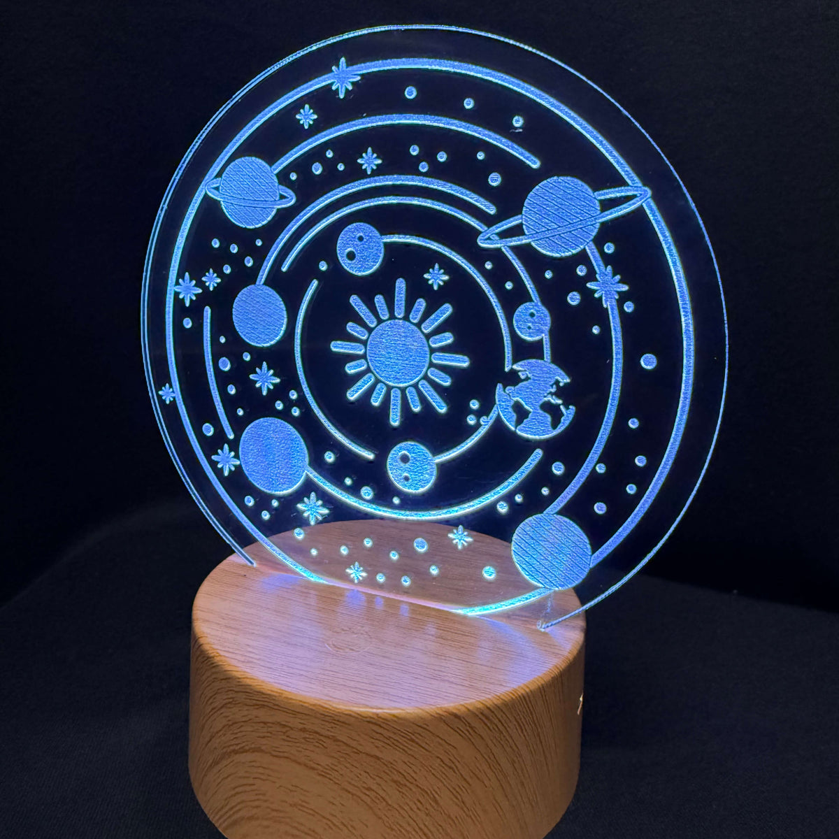 A 3D optical illusion night light featuring a glowing blue solar system design on a clear circular acrylic panel, mounted on a cylindrical wooden base against a black background.