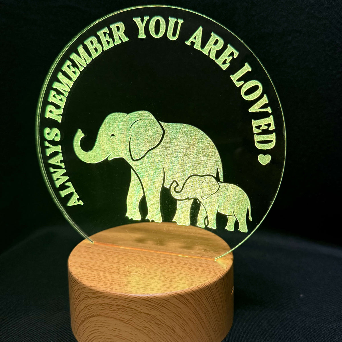 An acrylic lamp featuring a mother and baby elephant design with the text 'ALWAYS REMEMBER YOU ARE LOVED' in green, mounted on a cylindrical wooden base against a black background.