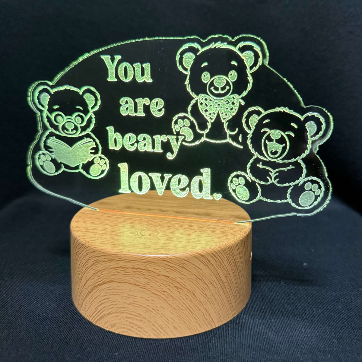 A wooden cylindrical base supporting a clear acrylic plaque engraved with teddy bear illustrations and the phrase 'You are beary loved.' in light green lettering, set against a dark fabric background.