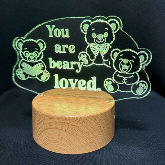 A wooden cylindrical base supporting a clear acrylic plaque engraved with teddy bear illustrations and the phrase 'You are beary loved.' in light green lettering, set against a dark fabric background.