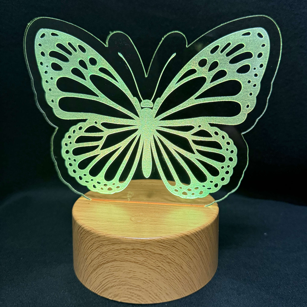 A butterfly-shaped LED light with a transparent cutout acrylic wing design resting on a round wooden base, set against a dark background.