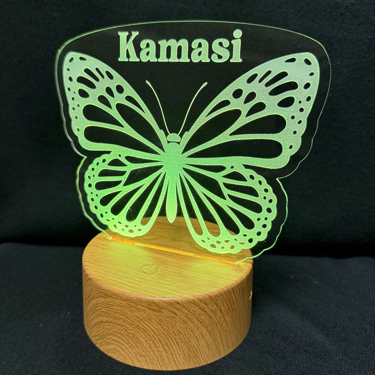 A personalized LED night light featuring a glowing green butterfly silhouette with the name 'Kamasi' cut out at the top, resting on a round, illuminated wooden base against a black background.