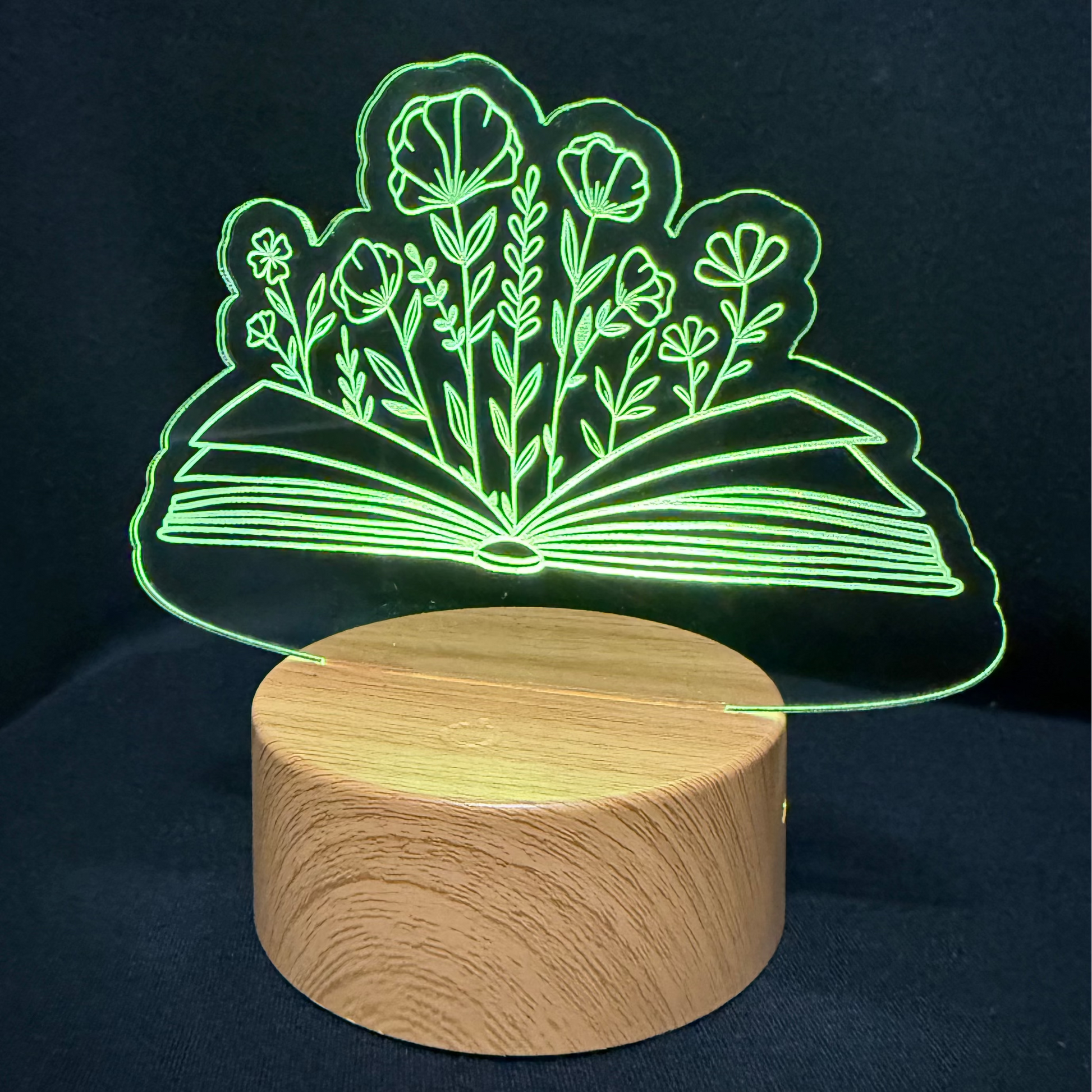 A 3D optical illusion night light featuring an open book with glowing green floral details, resting on a round, light wood-grain base against a dark background.