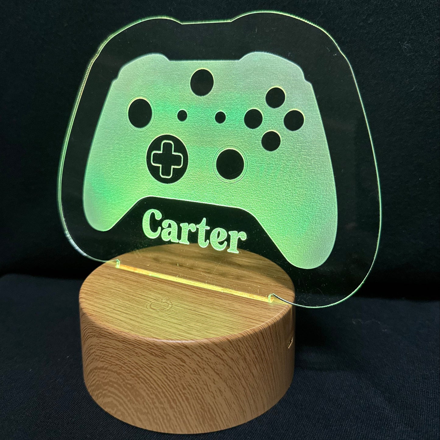 A personalized gaming-themed LED lamp featuring a translucent acrylic sign shaped like a game controller with the name 'Carter' engraved, illuminated in green, resting on a round wooden base against a black background.