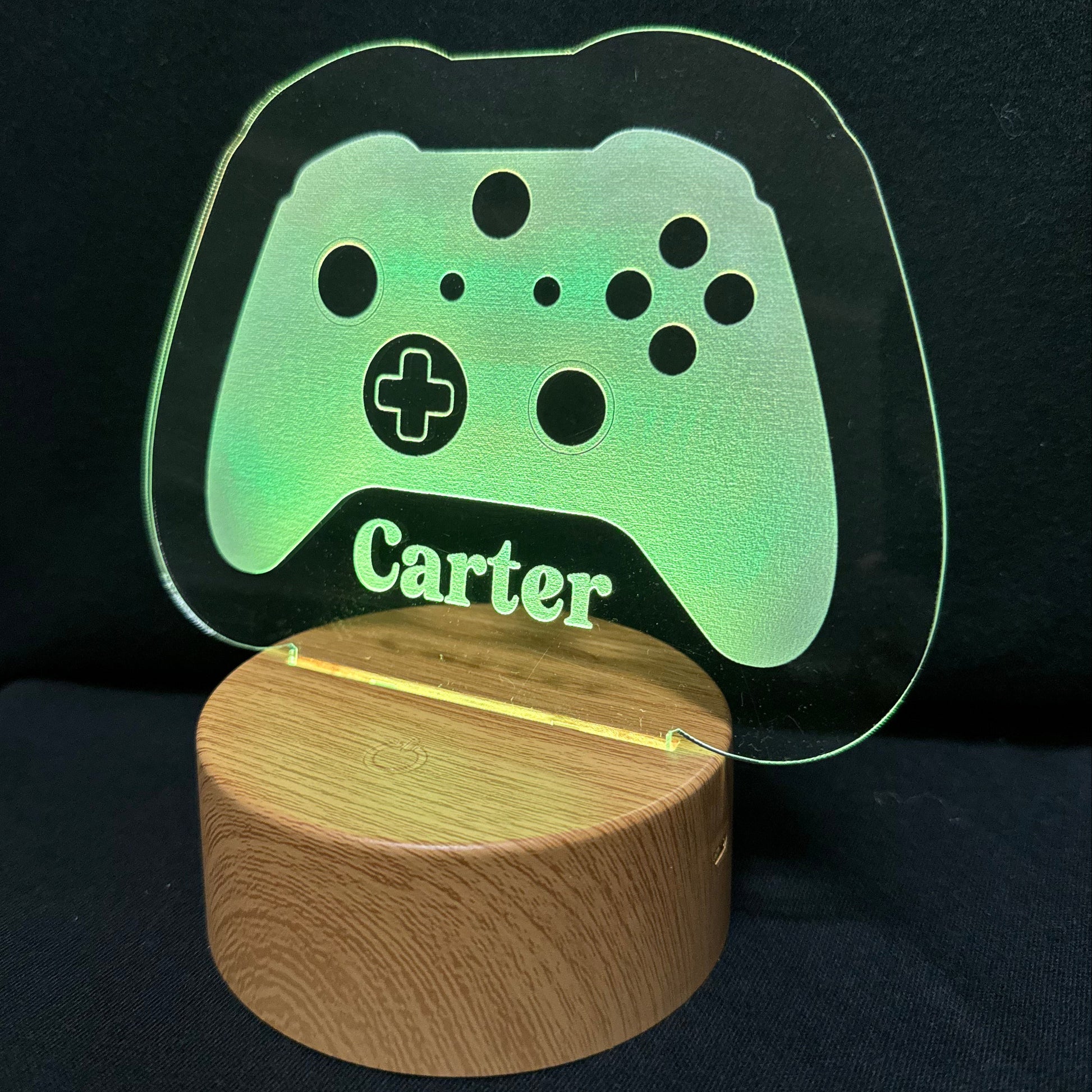 A personalized gaming-themed LED lamp featuring a translucent acrylic sign shaped like a game controller with the name 'Carter' engraved, illuminated in green, resting on a round wooden base against a black background.