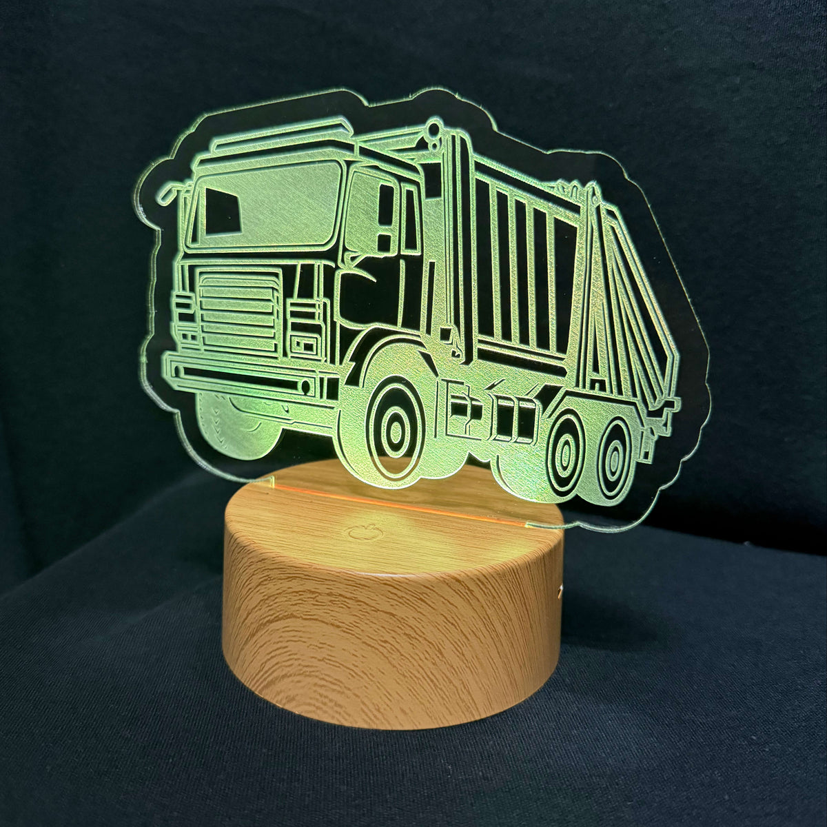 An LED acrylic night light featuring a glowing green garbage truck silhouette on a round wooden base, set against a dark background.