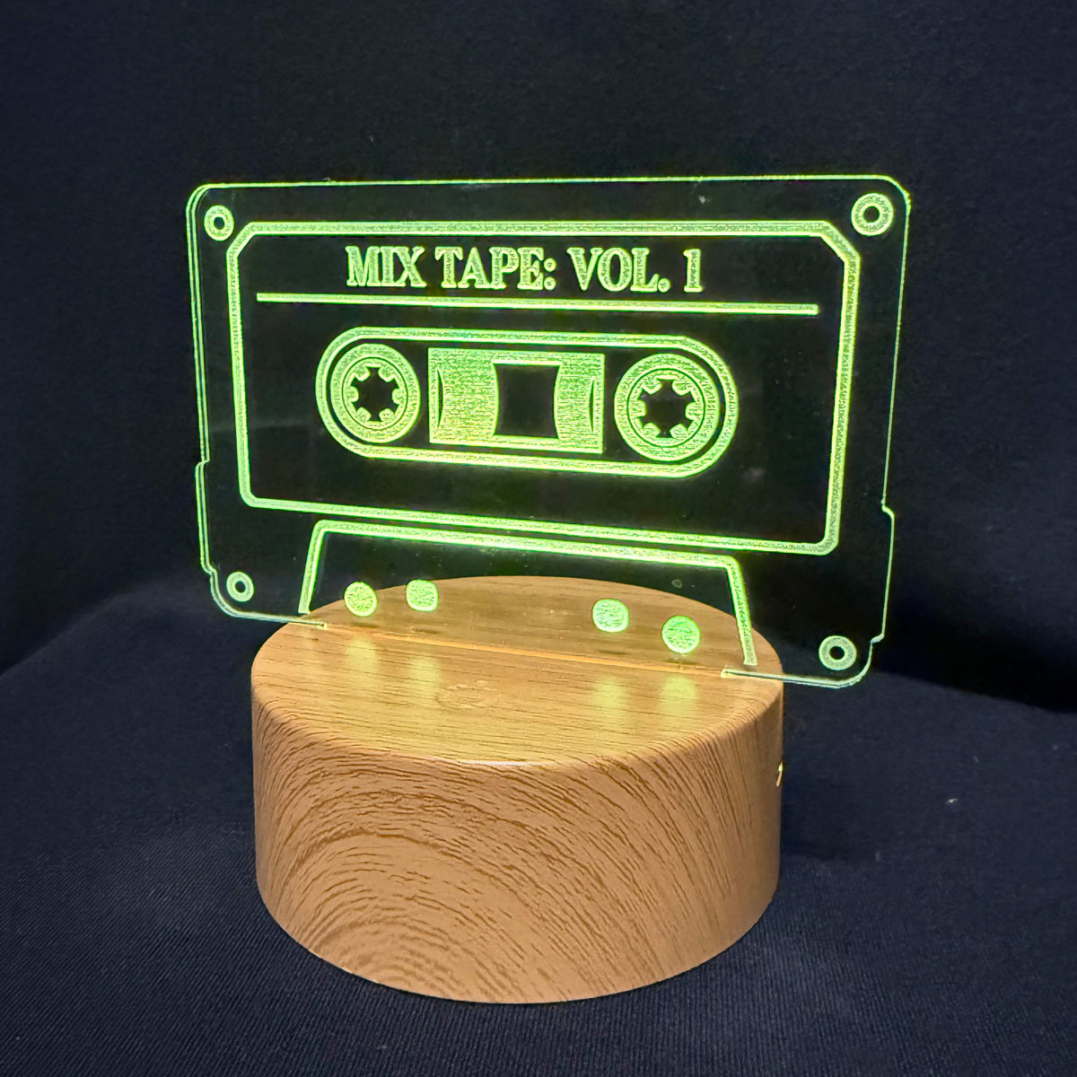 A cassette tape-shaped acrylic light on a round wooden base, glowing green against a dark navy background. The acrylic panel reads 'MIX TAPE: VOL.
