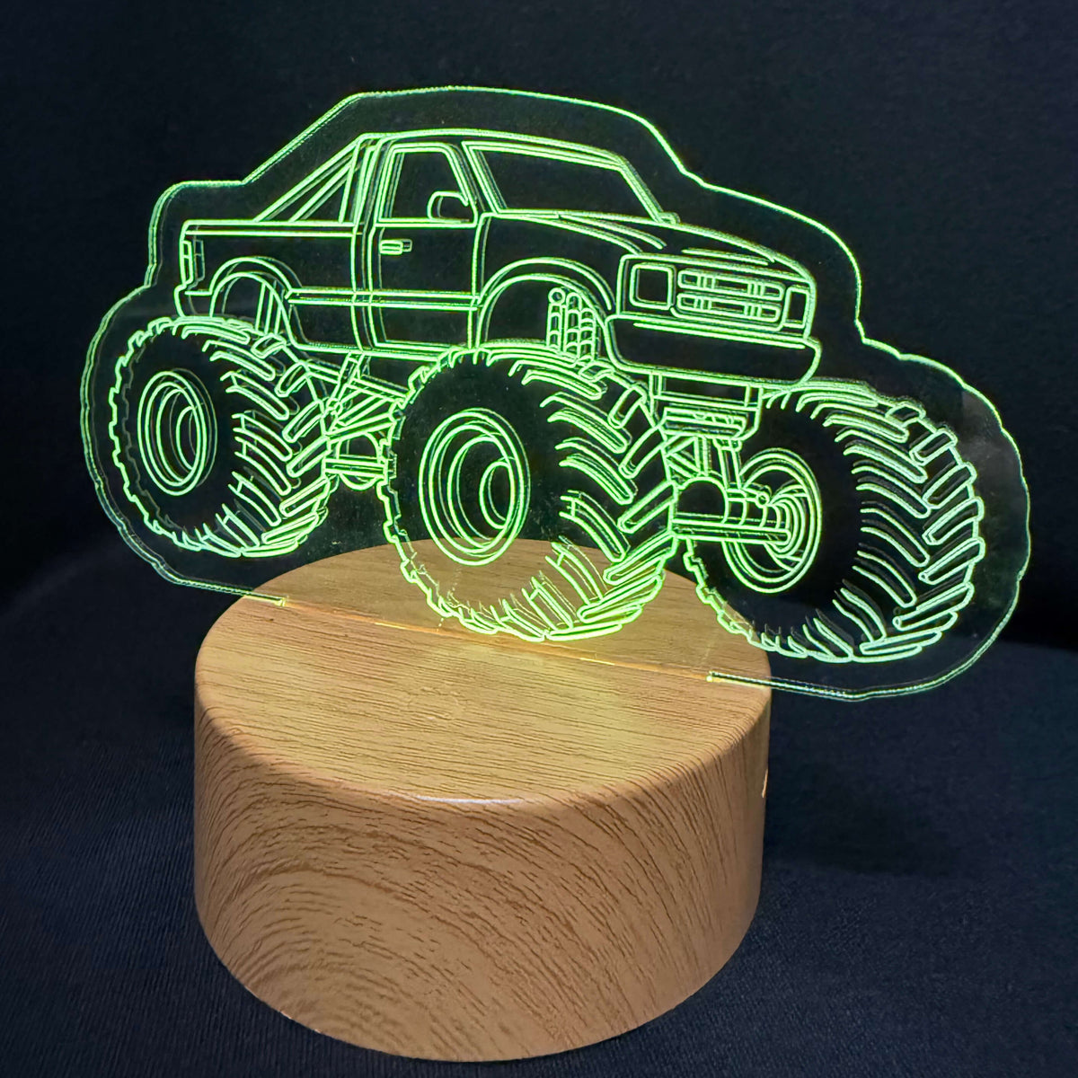 A 3D optical illusion night light featuring a glowing green outline of a monster truck on a round wooden base, set against a dark background.