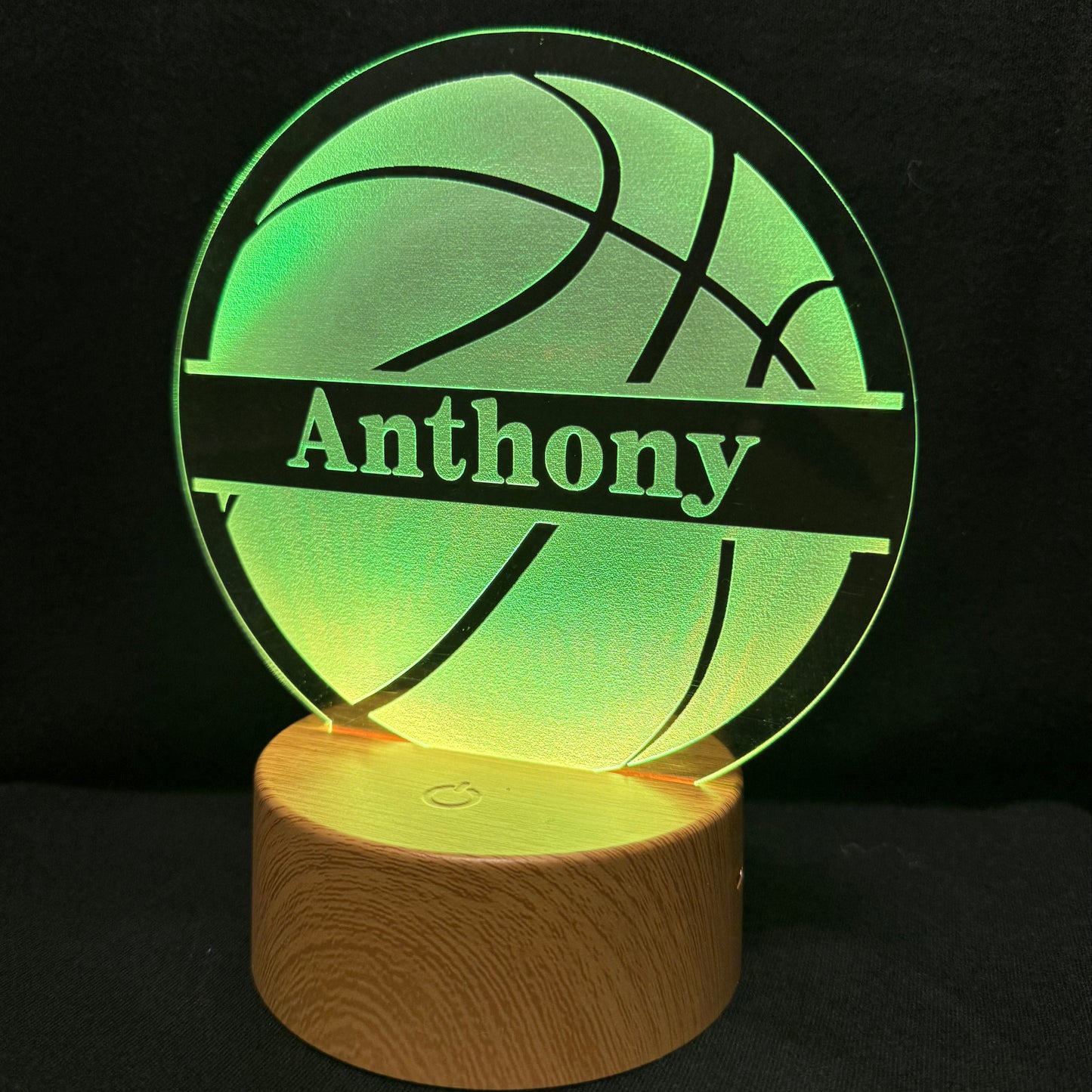 A personalized 3D optical illusion LED lamp featuring a basketball design with the name 'Anthony' engraved on a glowing green circular panel, mounted on a cylindrical wooden base against a black background.