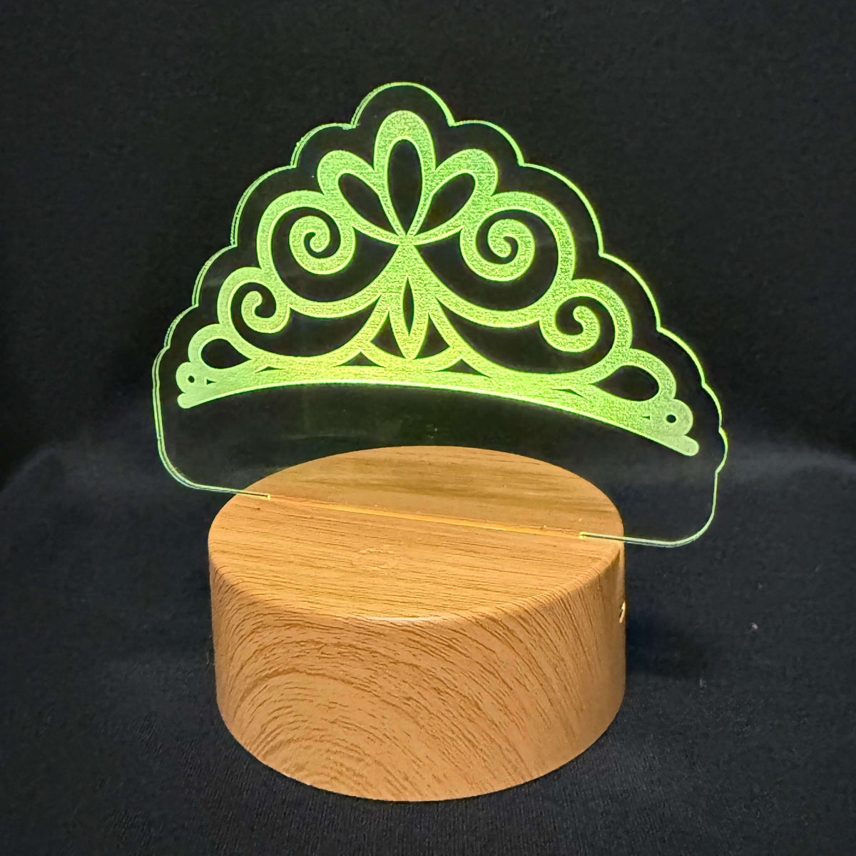 A clear acrylic tiara-shaped night light glowing green, mounted on a round light wood base against a black background.