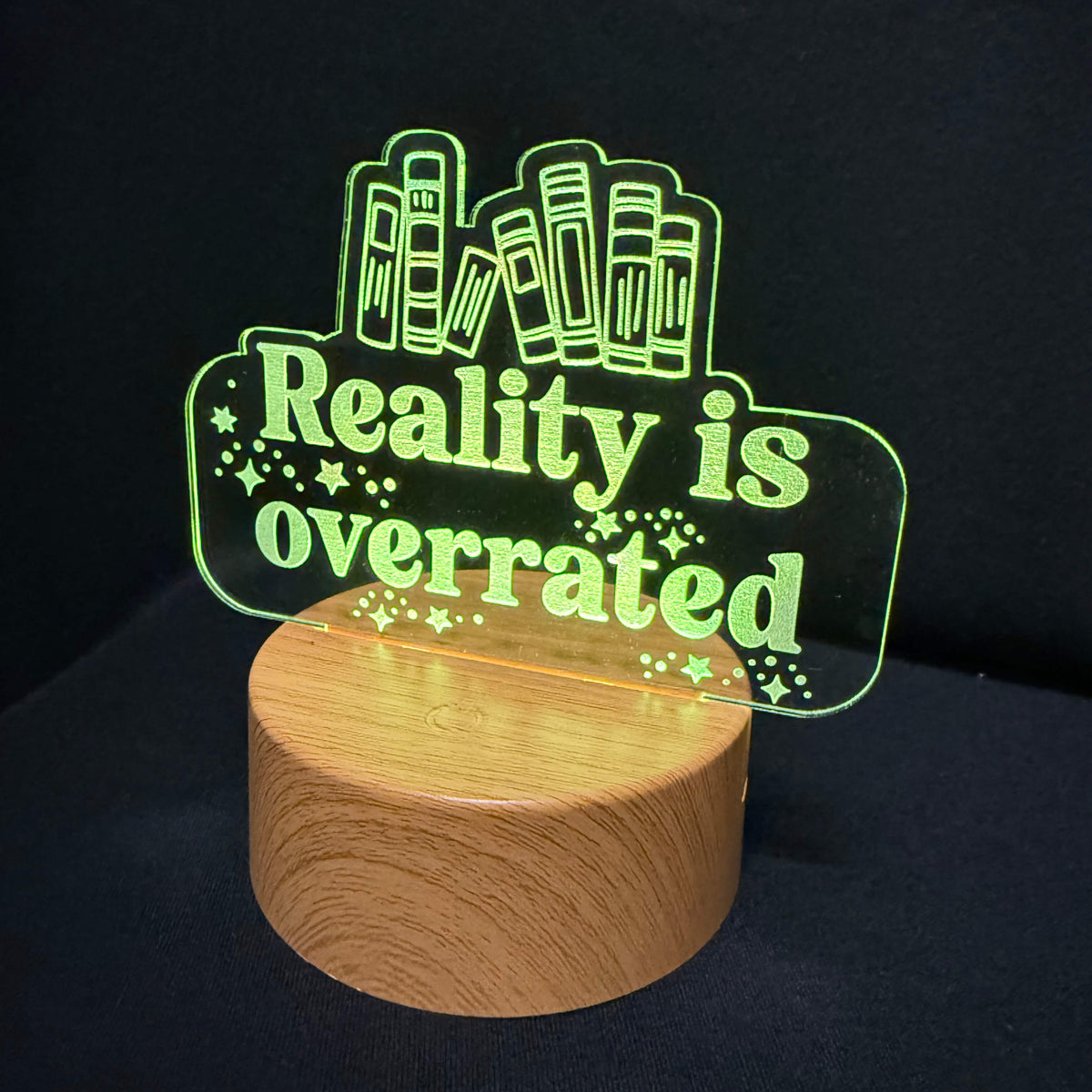 An LED acrylic night light with the text 'Reality is overrated' in glowing green, mounted on a cylindrical wooden base against a dark background.
