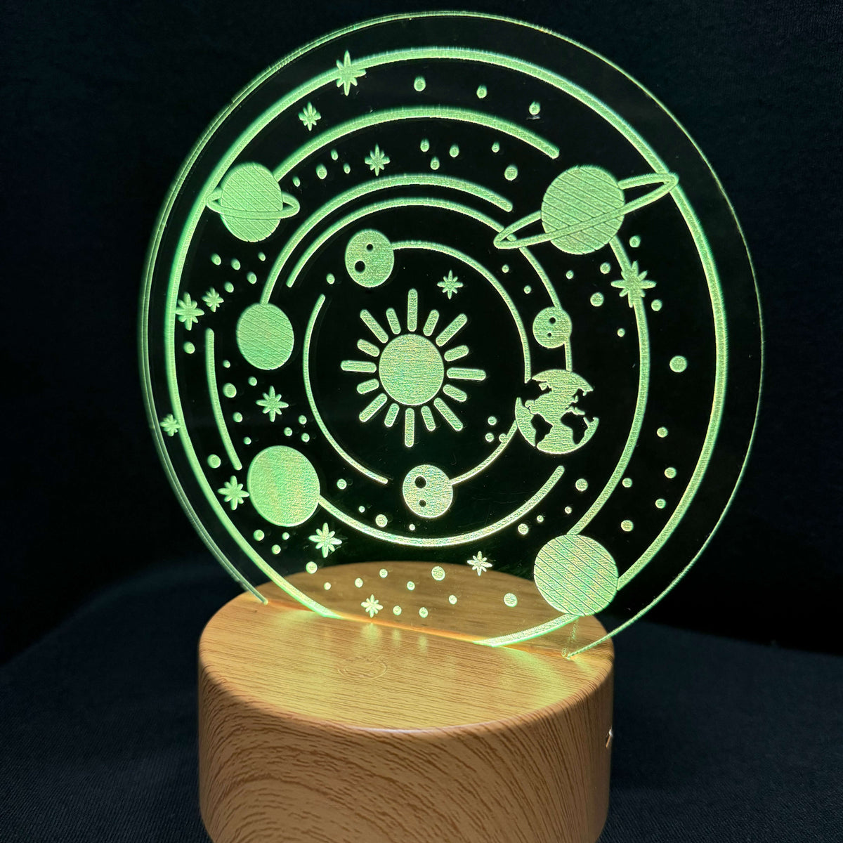A 3D optical illusion night light featuring a glowing green solar system design on a circular clear acrylic plate, mounted on a cylindrical wooden base against a black background.