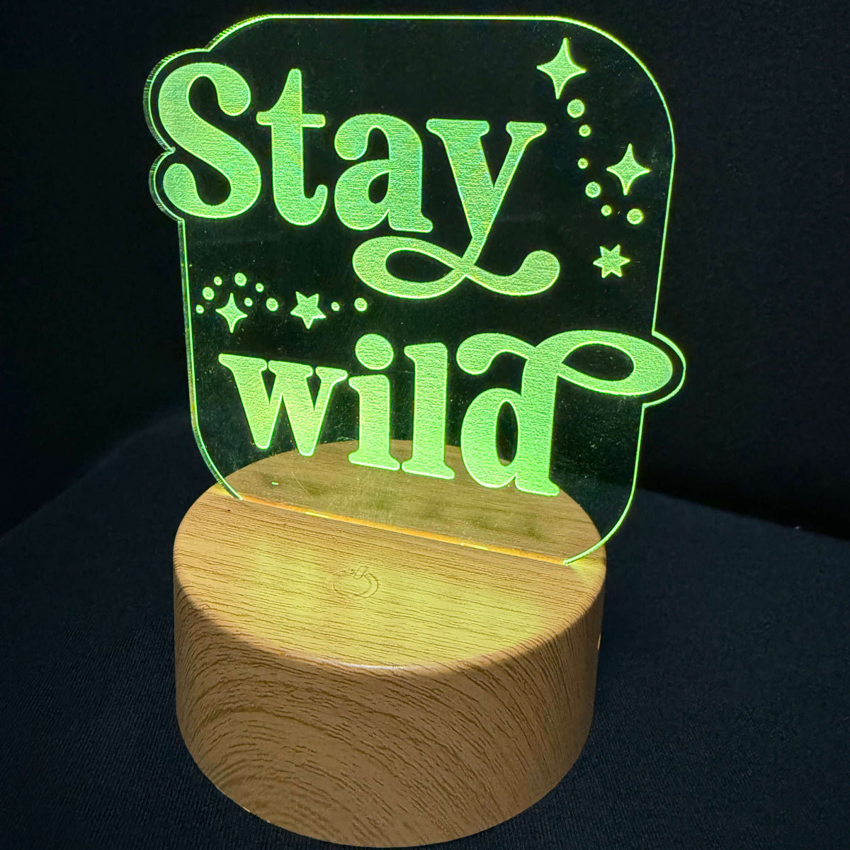 A 3D acrylic LED night light with the words 'Stay wild' in glowing green lettering, mounted on a round wooden base and photographed against a dark background.
